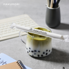 A glass mug of iced matcha milk tea with black tapioca pearls sits on a modern desk beside a notebook and keyboard. A wrapped Hakumori-branded paper straw rests across the rim.