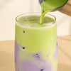 A close-up of a two-layer iced drink in a clear glass, featuring a vibrant purple taro slush base and a creamy green matcha latte top, being poured gently to create a marbled effect.